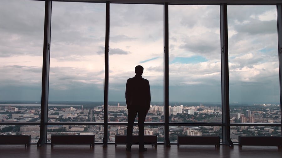 Silhouette of a man in a suit standing in front of floor-to-ceiling windows, overlooking a city skyline.