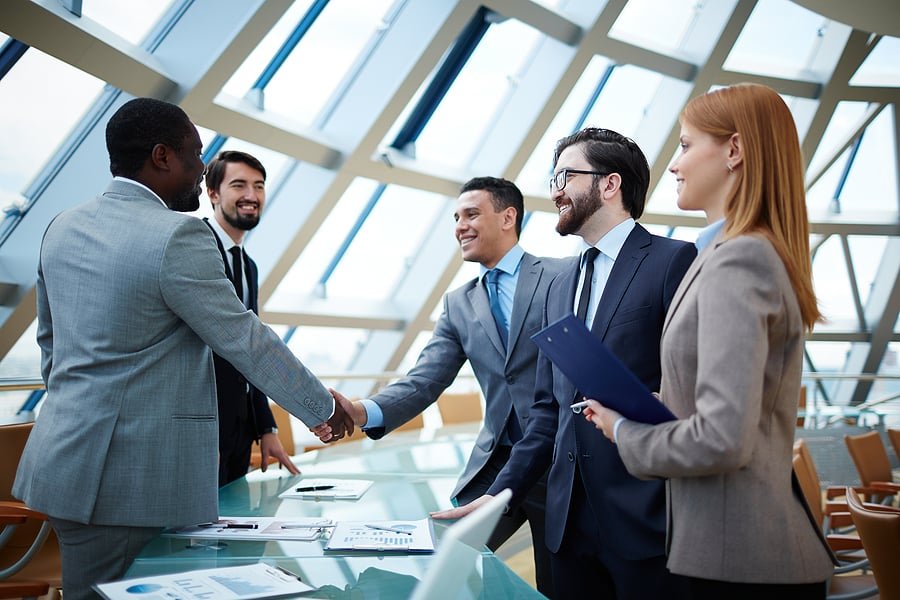 Group of professionals in business attire shake hands across a glass table in a bright, modern office.